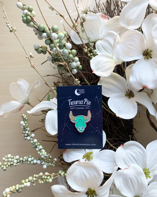Taurus Pin in Pastel Ming Green with Pastel Alternating horns with gold Enamel sitting on table with orchids.