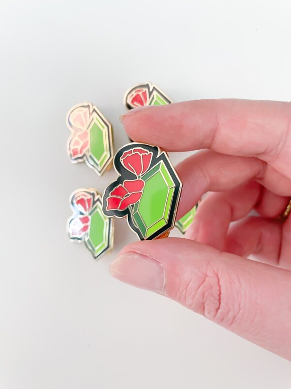 August Birth pin in hand featuring poppy flower and green peridot in gold enamel.